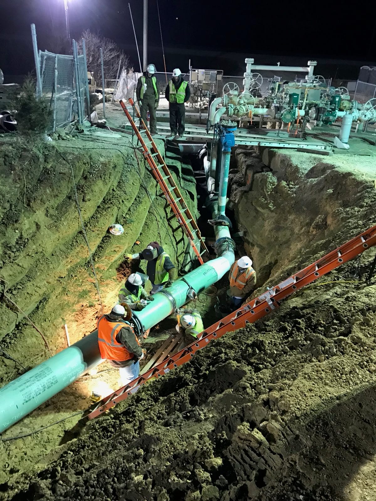 An Appalachian Pipeline crew in full PPE lowering coated pipe into a trench at a station tie-in at night